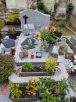 monument gris bleu moucheté avec stèle sur lquelle est fixée une croix noire à gauche, un espace jardinière en façade, de larges semelles avec de nombreuses plaques - O.Dufour