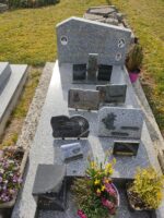 monument gris moucheté avec stèle sur laquelle figure les photos des défunts à côté de leur identification. En façade, dans l'espace jardiniere, il semblerait y avoir une urne funéraire avec les initiales JPM 6 o.Dufour